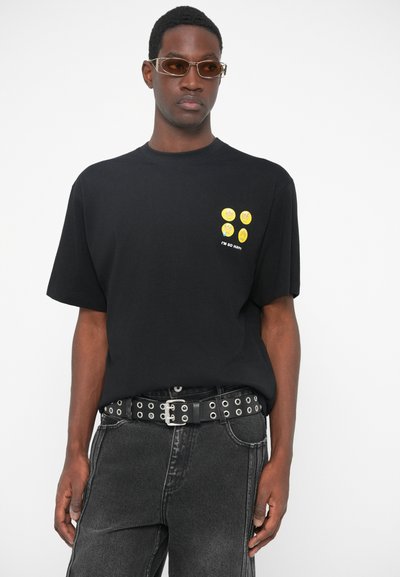 Sort kortærmet t-shirt med gule smiley-ansigt grafik på brystet, parret med mørkegrå jeans og et naglet sort bælte.