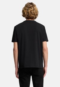 Czarna koszulka z krótkim rękawem i okrągłym dekoltem, o gładkiej fakturze i luźnym kroju, zestawiona z czarnymi spodniami, widziana od tyłu.