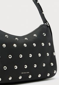 Μαύρη τσάντα ώμου από δέρμα με υφή πετρωμάτων, διακοσμημένη με ασημένια καρφιά, με καμπυλωτή σιλουέτα και έναν ρυθμιζόμενο λουρί. Εμφανίζεται το όνομα της μάρκας.