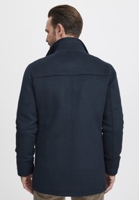 Mornarski vuneni kaput prikazan sa stražnje strane, s visokom ovratnikom, krojem koji ističe oblik tijela i vidljivim šavovima. Tekstura izgleda glatko i elegantno.