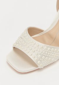 Λευκό σατέν σανδάλι με αγκράφα που διαθέτει φαρδιά λωρίδα διακοσμημένη με ποικιλία μεγεθών από στολιδάκια σε μορφή μαργαρίτη και τετράγωνο σχέδιο στη μύτη.