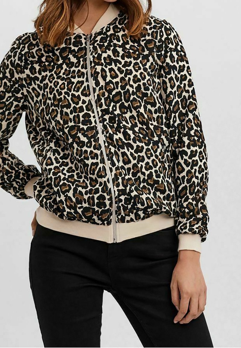 Femme portant une veste zippée à imprimé léopard avec des poignets et un col beige, associée à un pantalon noir, la main posée sur la hanche.