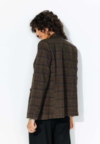 Blazer écossais marron en tissu texturé, présentant un design structuré, une couture d'épaule et des accents de boutons sur les poignets.