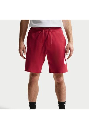 Hombre con pantalones cortos rojos con cordón y logo blanco en el muslo izquierdo, calcetines negros y camiseta blanca, de pie frente a un fondo liso.