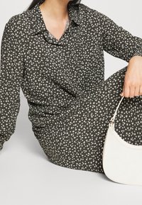 Blouse vert foncé avec un petit imprimé floral blanc, design à boutons et col. Sac à main blanc avec une forme incurvée et une bandoulière.