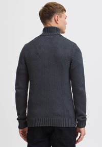 Mörkgrå stickad tröja med hög krage, ribbade muddar och nederkant. Har ett strukturerat mönster och en avslappnad passform.