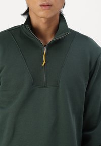 Sudadera de color verde oscuro con cremallera de un cuarto, hecha de un tejido suave, que presenta una textura acanalada en el cuello y un tirador de cremallera amarillo para contraste.