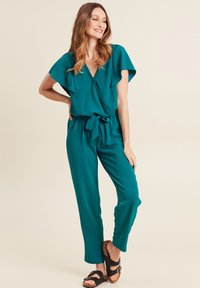 Tuta teal con maniche a sbuffo, scollatura a V, vita con fiocco e gamba dritta. Il modello indossa sandali neri. Tessuto liscio, leggero.