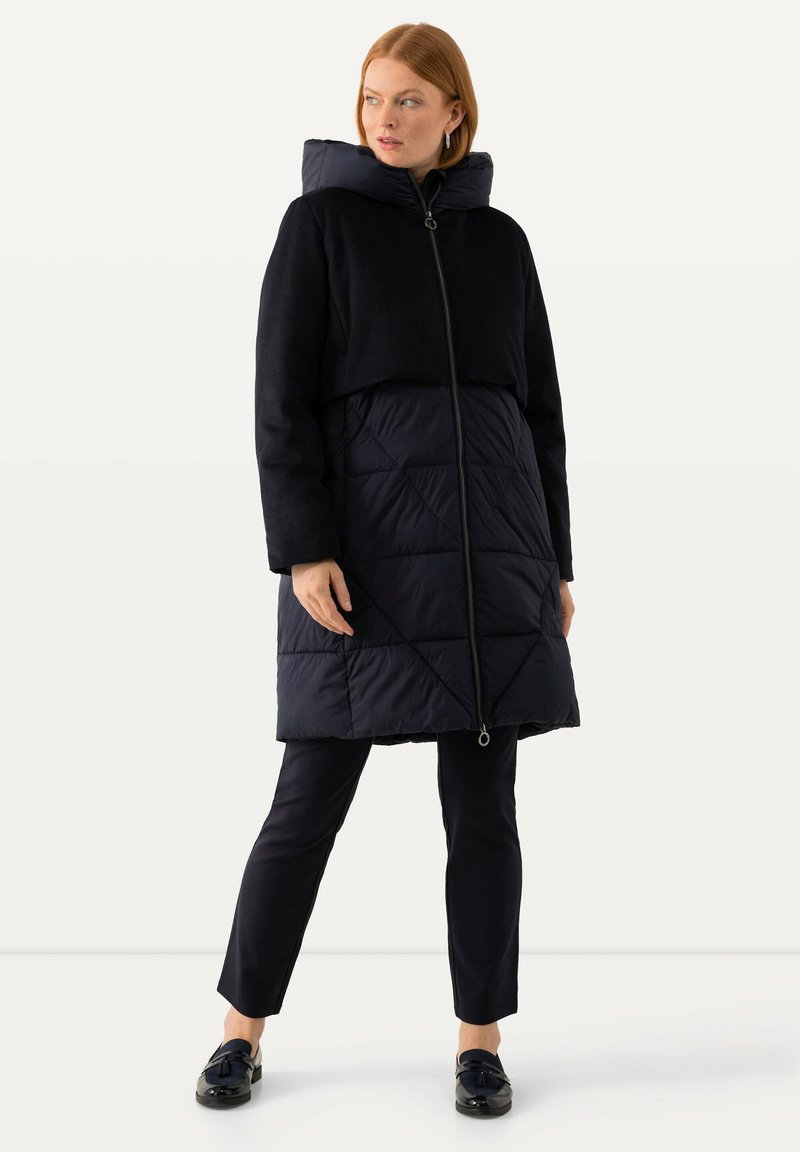 Svart pufferjacka med quiltad nederdel, fleeceöverdel och avtagbar huva. Har en framzipper och sidofickor.