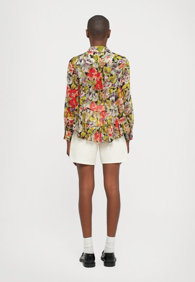 Bluză florală cu un model galben, roșu și verde, cu mâneci lungi și tiv ondulat, asociată cu pantaloni scurți albi și pantofi negri.