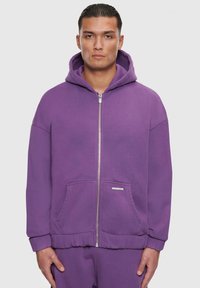 Lila Zip-Up Hoodie mit Kapuze, Fronttaschen, gerippten Bündchen und Saum. Hergestellt aus weichem Stoff mit glatter Textur und minimalem Branding.