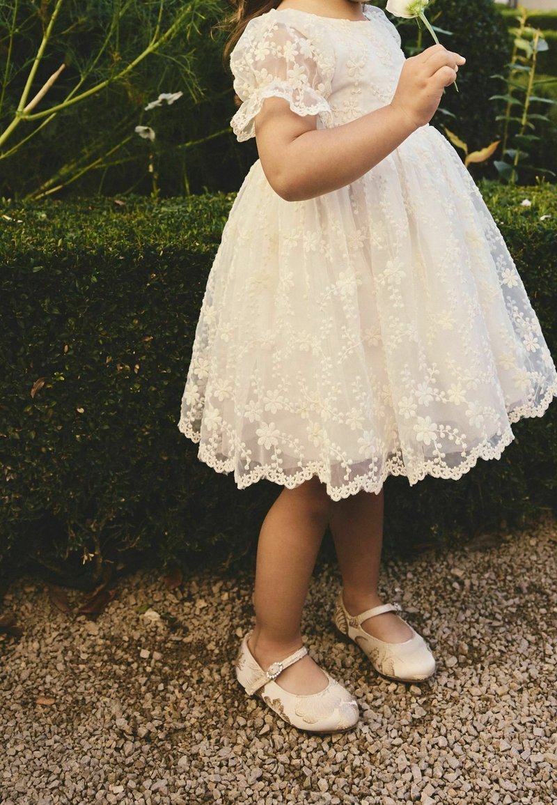 Jeune enfant en robe blanche en dentelle florale avec chaussures assorties tenant une fleur blanche, debout sur des graviers près de buissons verts taillés.
