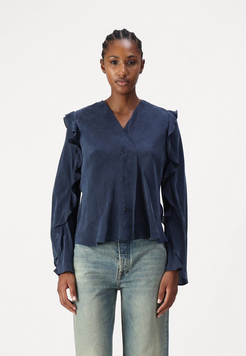 Navy blue blouse with ruffled sleeves and a V-neckline, featuring a button closure and textured fabric. Paired with light denim jeans.