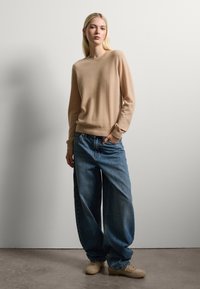 Maglione in maglia beige con scollatura rotonda, abbinato a jeans di denim blu oversize e scarpe beige. Texture liscia, vestibilità casual.