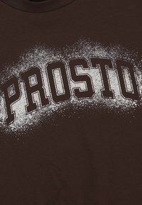 Brązowa bawełniana koszulka z napisem "PROSTO" w wyróżniających się białych literach, z teksturowanym, splamionym wzorem graficznym.