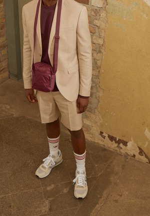 Hombre con un blazer beige y pantalones cortos, camisa burdeos, zapatillas blancas, calcetines a rayas y un bolso cruzado burdeos contra una pared texturizada.