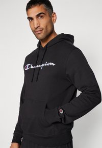 Hoodie noir avec une poche avant, capuche avec cordon de serrage, et logo "Champion" brodé en blanc et rouge. Texture douce, coupe décontractée.