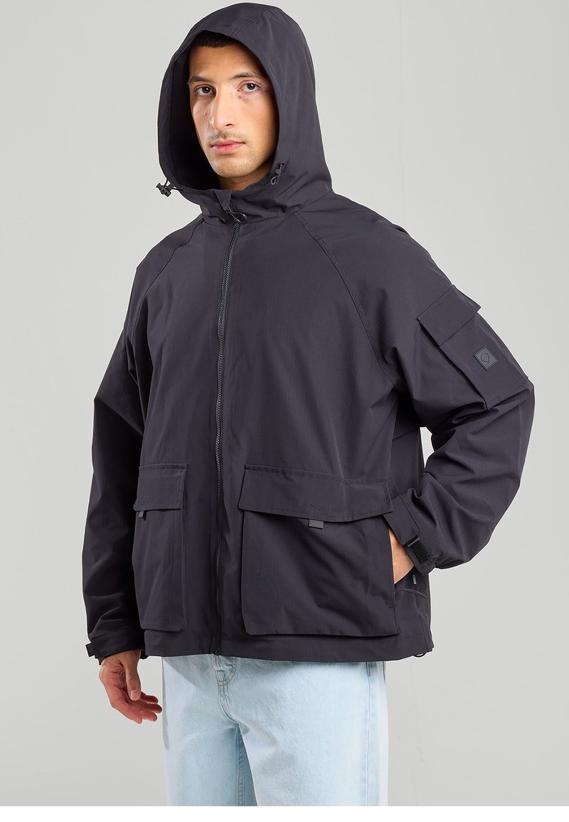 Black hooded jacket featuring a front zipper, two large front pockets, and adjustable cuffs. Made of lightweight, water-resistant fabric.