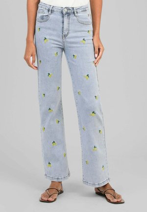 Jean en denim bleu clair avec une petite broderie jaune citron, porté avec des sandales marron à lanières sur un fond neutre.