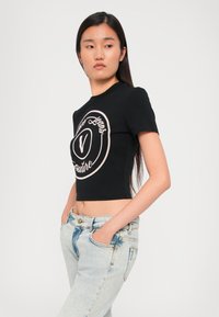 Juodas trumpas marškinėlis su trumpomis rankovėmis, turintis rožinį apvalų logotipą. Derinami su šviesiai mėlynomis džinsinėmis kelnėmis, perteikiančiomis laisvą siluetą.