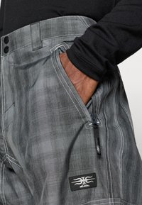 Sive vzorirane cargo hlače z belimi šivi, ki imajo stranski žep in majhno označevalno nalepko blagovne znamke spredaj.