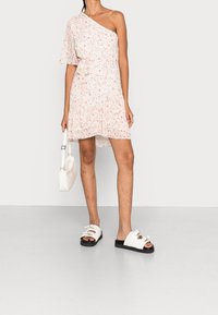 Jednoskrzydłowa sukienka w kwiaty w jasnoróżowym kolorze z małymi, wielokolorowymi kwiatkami, związana w talii, połączona z białymi sandałami i białą torebką.