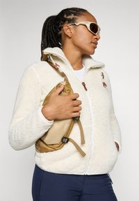 Veste en polaire blanche avec un col haut, des accents marron et une fermeture éclair à l'avant. Tient un sac kaki avec des bretelles réglables. Pantalons sombres visibles.