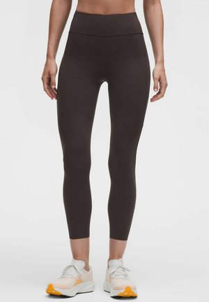 Kvinde iført høj talje mørkebrune leggings og hvide sneakers med orange såler, stående mod en ensfarvet lys baggrund.