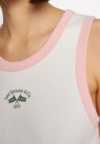 Vit tanktopp med rosa ribbad rund halsringning, prydd med en grön logotyp med korsade flaggor och texten "Levi Strauss & Co. 1873" över bröstet.