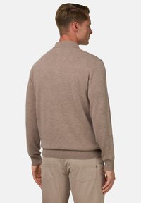 Švarkelis šviesiai rudos spalvos su trikotažiniu apykaklė, raukšlėtais rankogaliais ir apdaila; dėvimas su šviesiai smėlio spalvos kelnėmis; lygus paviršius, kasdienis dizainas.