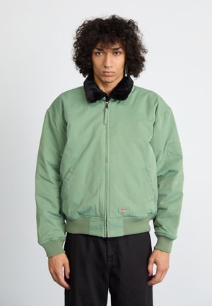 Chaqueta bomber verde, fabricada con tejido acolchado, con cuello de piel sintética negra, cierre frontal, dos bolsillos laterales, puños y dobladillo acanalados, parche de la marca en la parte inferior izquierda.