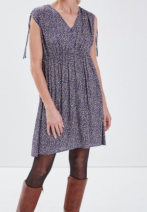 Kobieta ubrana w krótką granatową sukienkę w kwiaty z krótkimi rękawami, czarne rajstopy i brązowe kozaki za kolano na jednolitym białym tle.