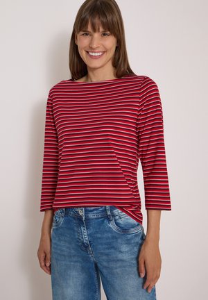 Femme portant un haut à manches longues rayé rouge et blanc avec des manches trois-quarts et un jean bleu en denim, souriant à la caméra.