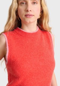 Tajta ärmlös stickad topp i klar korall med en mjuk, fluffig textur. Har hög krage och kontrasterande ljusrosa kanter.