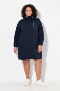 Krøllet kvinde iført en navyblå langærmet hættetrøjedragt med snore og hvide sneakers, stående mod en hvid baggrund.