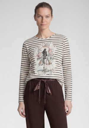 Frau trägt ein gestreiftes Langarmshirt mit einer Modeillustration und dem Text "Pretty Things," kombiniert mit dunklen Hosen und einem gebundenen Gürtel.