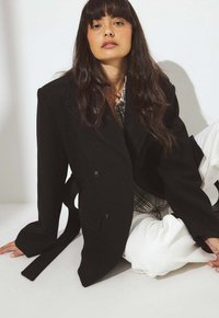 Must topelt-rinnaga mantel suurte kraedega, tekstureeritud kangast, külgedel lipsud ja nupuga kaunistused. Kandmise all ruuduline särk ja valged püksid.