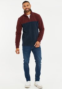 Pullover a mezzo zip in pile bordeaux e blu navy, caratterizzato da una finitura testurizzata e logo ricamato sul petto. Indossato con jeans e sneakers.