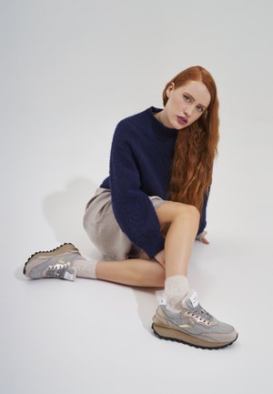 Sportowe sneakersy w odcieniach szaro-beżowych z teksturowanym materiałem, kontrastującymi złotymi akcentami oraz masywną czarną podeszwą, w zestawie z jasnymi skarpetkami.