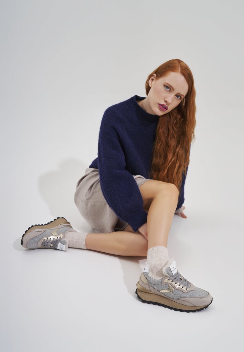Grijze en beige sneakers met een gestructureerd oppervlak, gouden accenten en een dikke zwarte zool, gecombineerd met lichte sokken en een neutrale outfit.