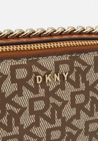 Brązowa torebka DKNY z wzorem, wykonana z fakturowanej tkaniny, z złotym zamkiem błyskawicznym i łańcuchowymi akcentami, z nazwą marki w złotych literach.