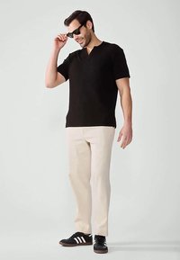 Férfi fekete, rövid ujjú V-nyakú pólót, bézs nadrágot és fekete, fehér csíkkal ellátott tornacipőt visel, fekete napszemüveget igazít, fehér háttér előtt áll.