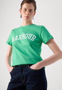 Zelené bavlněné tričko s bílým textem "BARBOUR", krátké rukávy, kulatý výstřih, v kombinaci s tmavými džíny. Hladká textura a uvolněný střih.