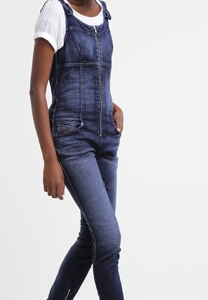Femme portant une salopette en denim bleu foncé avec fermeture éclair et poches avant, sur un t-shirt blanc à manches courtes, debout devant un fond clair uni.