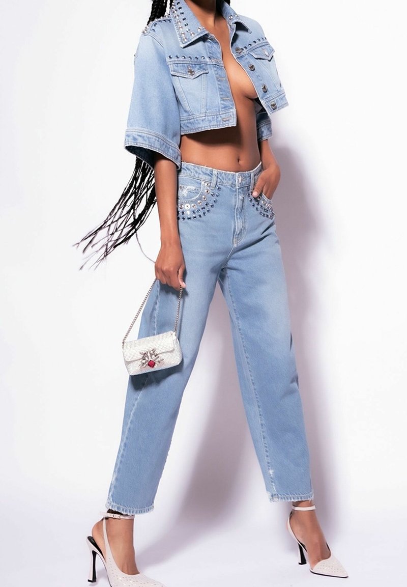 Veste en denim bleu clair raccourcie avec des clous argentés, associée à un jean bleu clair taille haute avec des accents cloutés. Le mannequin tient un sac à main blanc.
