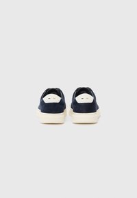 Sapatos de camurça azul-marinho sem atacadores, com um detalhe branco no calcanhar e uma sola de borracha creme, apresentando uma textura suave e um design minimalista.