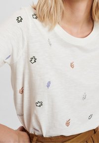 Kobieta ubrana w białą koszulę z krótkim rękawem, haftowaną w małe zielone, fioletowe i brązowe liście, wpuszczoną w brązowe spodnie.