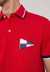 Polo shirt rossa con colletto a contrasto a righe blu e bianche, e un patch triangolare con cuciture blu, bianche e rosse.
