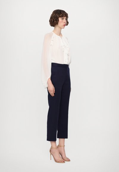 White blouse with ruffles, navy tailored trousers, and nude strappy heels. Model stands side-on, showcasing the outfit's silhouette.