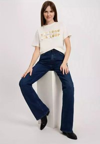 Biała koszulka z złotym napisem, zestawiona z ciemnoniebieskimi spodniami dzwonami. Modelka stoi na białym piedestale, mając na sobie czarne botki sięgające do kostki.